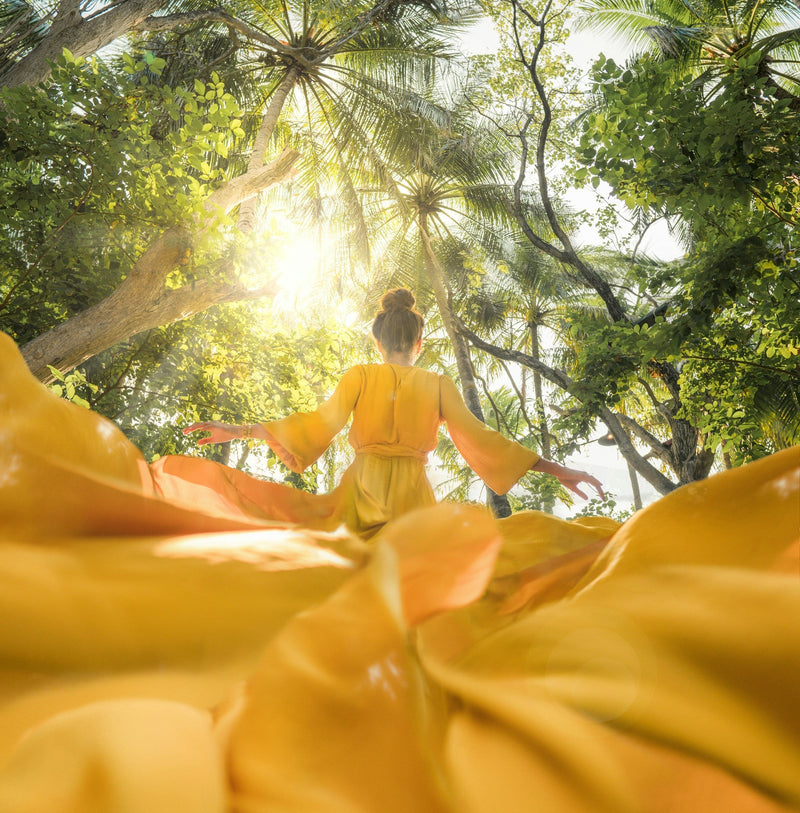 picture of a woman from behind wearing a billowing golden yellow dress. the picture is taken from below so you see the tree canopy above her with the sun shining through.