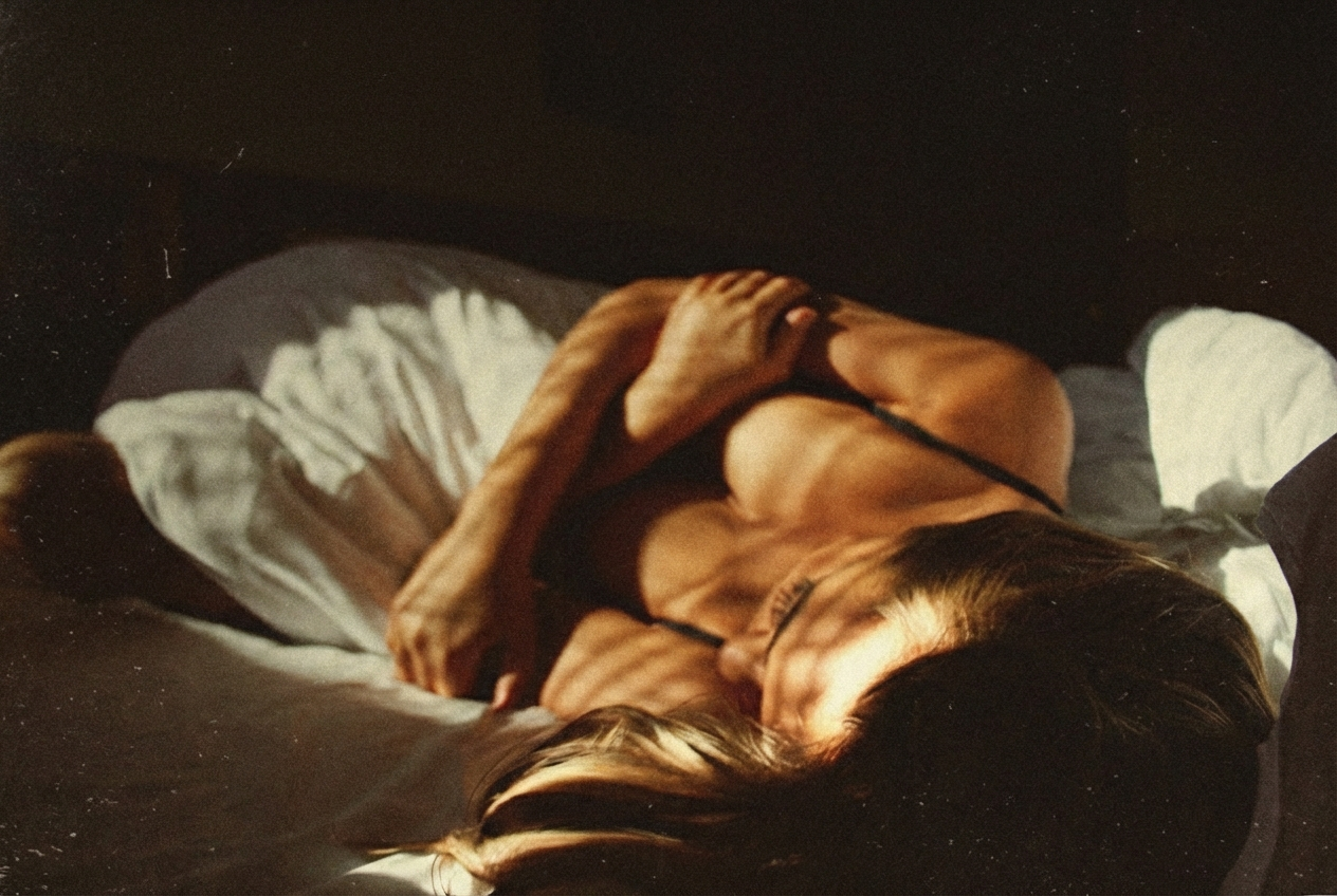 Warm, cinematic photograph of a woman lying curled on white sheets, sunlight casting soft striped shadows across her face and bare shoulders, creating an intimate, golden-hour atmosphere.